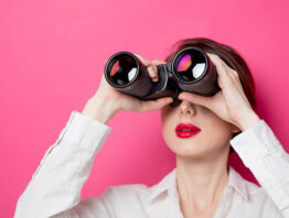Portrait of young businesswoman with binoculars on pink background.