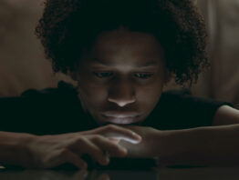 Boy leaning on table staring at smartphone, illuminated by screen light, distant expression, emotional isolation and apathy in digital age captured